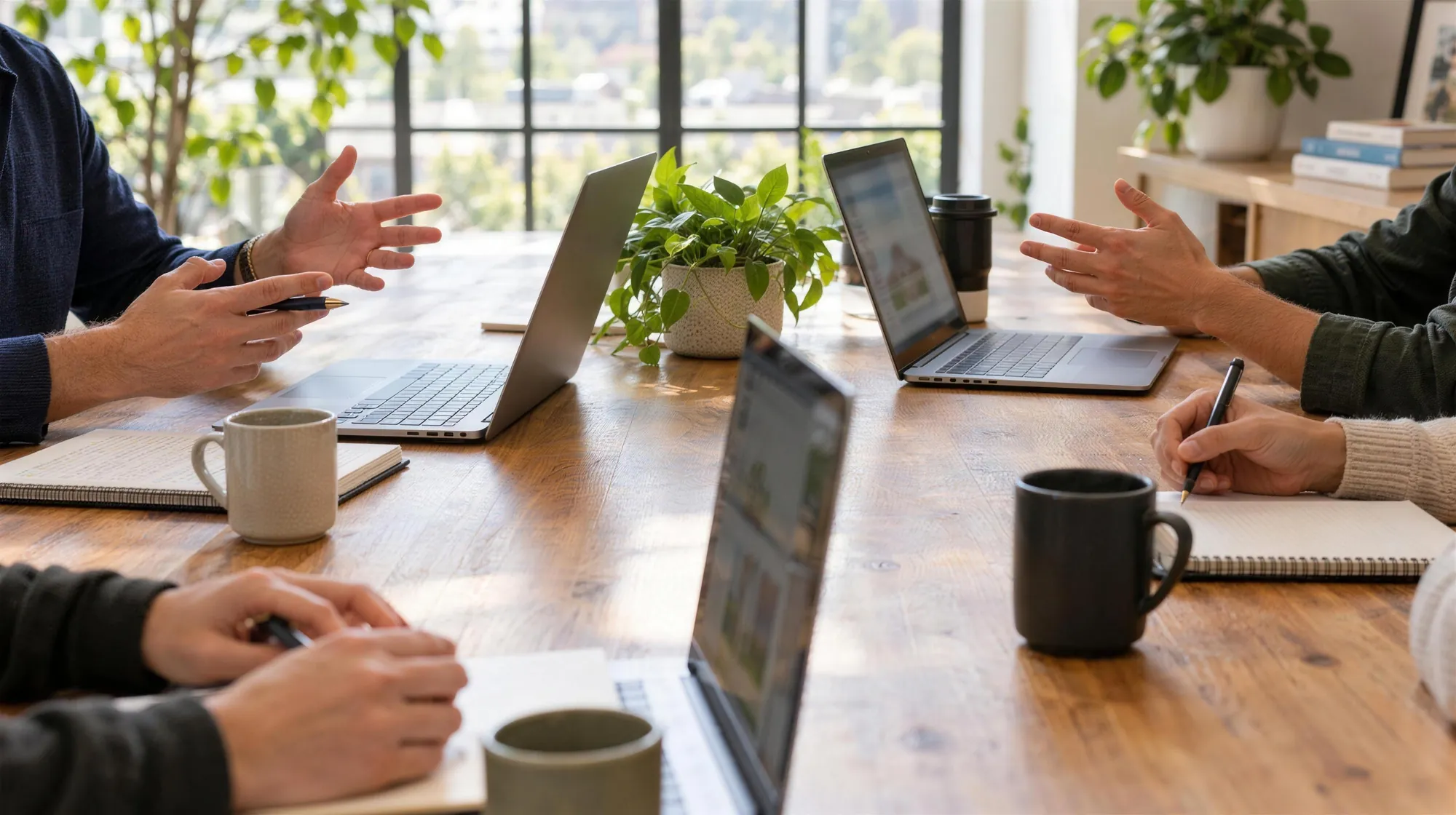 A small real estate team working around a long wooden table in a bright office, laptops and phones out, one agent on a call by the window